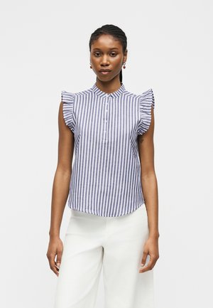 Kobieta w niebiesko-białej pionowo paskowanej bezrękawnikowej bluzce z falbankami na ramionach i białych spodniach, stojąca na jednolitym tle.