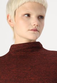 Pulover roșu texturat cu guler înalt, expus pe un model, prezentând o vedere de aproape a decolteului și feței.