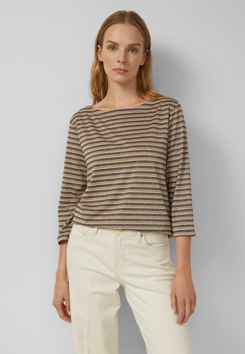 Frau mit langen blonden Haaren, die ein beigefarbenes und braun gestreiftes Langarmshirt und off-white Hosen trägt, steht vor einer grauen Wand.