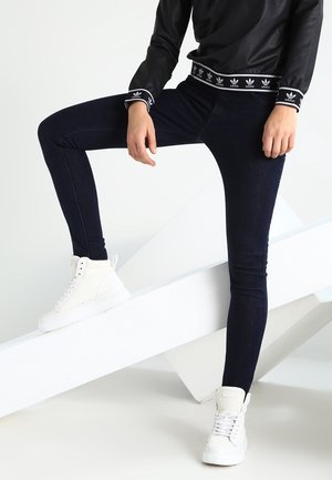 Personne portant un haut Adidas noir, un jean skinny foncé et des baskets montantes blanches, posant un pied sur une surface blanche inclinée.