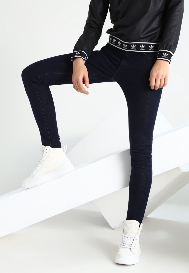Personne portant un haut Adidas noir, un jean skinny foncé et des baskets montantes blanches, posant un pied sur une surface blanche inclinée.