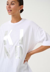 T-shirt oversized blanc avec des lettres métalliques argentées qui écrivent "KIND." Le tissu est lisse avec une coupe décontractée et des manches courtes.