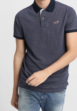 Marineblå poloshirt med tekstureret stof, kontrasterende krave og ærmekanter, prydet med et lille broderet logo på brystet.