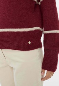 Bordeaux gebreide trui met crèmekleurige strepen, geribbelde manchetten, gestructureerde afwerking en een klein metallic logo detail op de zoom. Gestyled met beige corduroy broek.