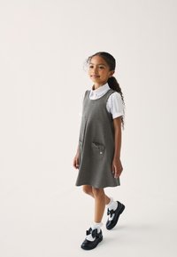 Graues Kleid mit ärmellosem Design, das zwei Fronttaschen mit Schleifendetails aufweist. Kombiniert mit schwarzen Schuhen und gerüschten weißen Socken.