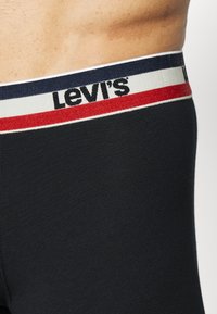 Melna apakšveļa ar baltu jostu, uz kuras redzama sarkana un tumši zila svītra, kā arī Levi's logo izcili attēlots melnā krāsā.