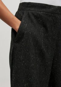 Schwarze Hose aus einem strukturierten Stoff mit dezenten Sprenkeln. Eine Seitenasche ist sichtbar, in der eine Hand ruht.