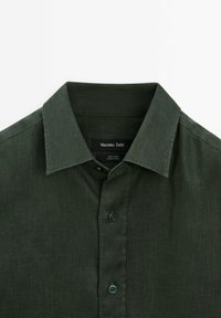 Chemise en lin vert foncé avec un col boutonné, dotée d'un tissu texturé et de deux boutons foncés à l'avant.