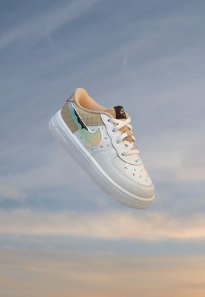Biela teniska Nike s viacfarebnými škvrnami, béžovou vnútornou podšívkou a irizujúcim logom, vznášajúca sa na modrom nebi.