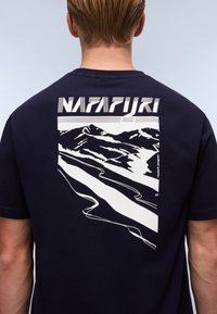 Granatowa koszulka T-shirt z dużym białym nadrukiem gór i wijącej się drogi na plecach, z napisem "NAPAPIJRI" powyżej.