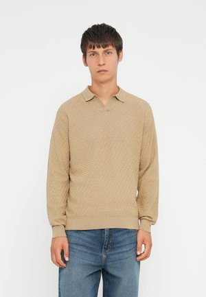Beige knitted pullover with a v-neck and collar. Textured pattern throughout, long sleeves, and ribbed cuffs and hem. Worn with blue jeans.
