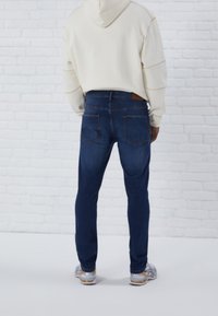Jeans de denim azul oscuro con corte ajustado, que cuentan con dos bolsillos traseros y una etiqueta de cuero marrón en la cinturilla. Llevados con una sudadera blanca.