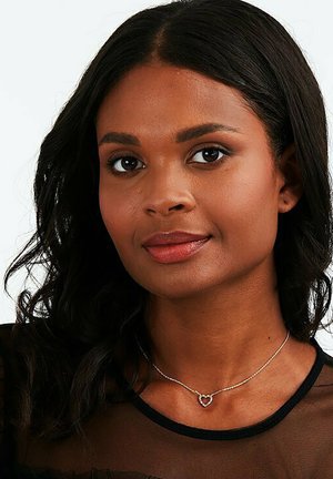 Silberne Halskette mit einem kleinen Herzanhänger an einer zarten Kette. Der Anhänger weist subtile Details auf und hebt sich vor einem schwarzen Oberteil ab.