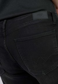 Jean en denim noir avec une finition texturée, une grande poche arrière avec une couture décorative et une étiquette en cuir marquée au niveau de la taille.