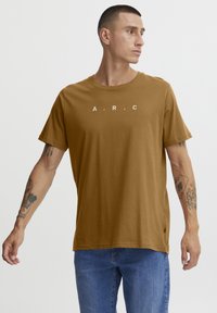 Brun bomull t-skjorte med korte ermer, rund hals og trykt "A.R.C." tekst i hvitt over brystet. Bæres sammen med blå jeans.