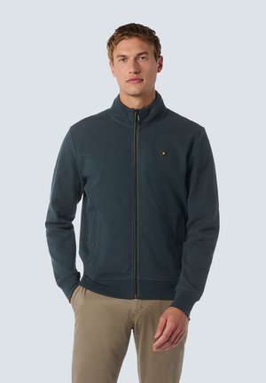 Donkere blauwe ritsjack met hoge kraag, zachte textuur, voorzakken en een klein logo op de borst. Draagt het met beige broeken.