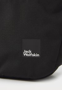 Svart stoffveske med teksturert overflate og firkantet logo-patch med "Jack Wolfskin" i hvit tekst. Enkel og funksjonell design.