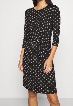 Femme portant une robe noire mi-longue aux manches trois quarts, ornée d'un petit motif de fleurs blanches, avec une ceinture nouée à la taille.