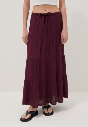Femme portant une jupe bordeaux longue jusqu'au sol, à volants, avec une ceinture élastique et un cordon, associée à des sandales plateformes noires.