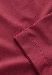 Burgundijas kokvilnas audums ar gludu tekstūru un redzamām šuvēm. Attēls parāda saliektu piedurkni, izceļot tās bezšuvju dizainu.