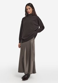 Ciemnobrązowy sweter z golfem w połączeniu z długą, lejącą się, błyszczącą szarą spódnicą z satyny. Modelka ma na sobie gładkie czarne botki za kostkę. Prosty, nowoczesny fason.