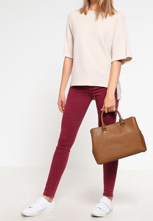 Femme portant un haut beige côtelé à manches courtes, un jean skinny bordeaux, des baskets blanches à enfiler, tenant un sac à main en cuir marron par ses anses.