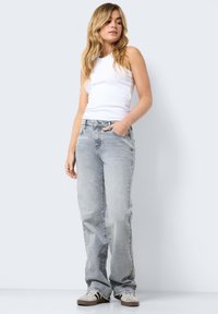 Jovem mulher com cabelo loiro longo, vestindo uma blusa branca sem mangas, jeans cinza claro e sapatilhas brancas com riscas pretas.