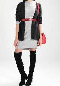 Abito grigio a maglia con cardigan nero textured, cintura rossa e borsa rossa. Stivali alti neri completano l'outfit.