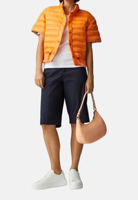 Osoba oblečená v oranžové krátké puffer bundě, bílé košili, tmavě modrých šortkách po kolena, bílých teniskách a držící béžovou kabelku.