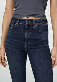 Jean bleu foncé taille haute avec une coupe ajustée, fabriqué en tissu denim. Comprend des poches avant et une petite fermeture à bouton métallique.