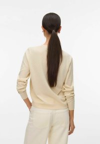 Pull beige en tricot avec un motif texturé, manches longues et bord-côtes, associé à un pantalon de couleur claire. Cheveux attachés en queue de cheval.