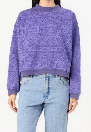 Osoba w fioletowej, wzorzystej bluzie z długim rękawem i jasnoniebieskich, wysokich spodniach jeansowych, stojąca na jednolitym tle.