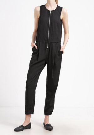 Combinaison noire sans manches avec fermeture éclair à l'avant, poches latérales, jambes fuselées, portée avec des chaussures noires texturées sans lacets sur un mannequin.