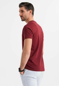 Bordó tričko s krátkým rukávem a kulatým výstřihem, slim fit, s hladkou bavlněnou texturou, nošené mužem stojícím bokem proti jednobarevnému pozadí.