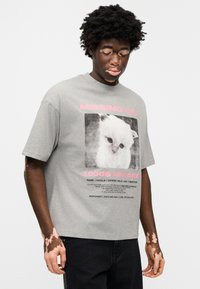 Γκρι βαμβακερό T-shirt με μπροστινό γραφικό ενός λευκού γάτου, περιτριγυρισμένο από ροζ μήνυμα "ΧΑΘΗΚΕ ΓΑΤΑ" και στοιχεία επικοινωνίας σε μαύρο κείμενο.