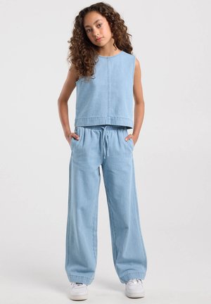 Persona joven con cabello rizado que lleva una camiseta sin mangas de mezclilla azul claro y pantalones anchos a juego con zapatillas blancas, manos en los bolsillos.