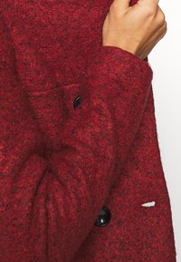 Rød uldfrakke med tekstureret finish, udstyret med en sort knap og en lomme. Stoffet virker tykt og varmt med en let frynset detalje.