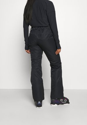 Henkilö, jolla on mustat hiihto- tai lumihousut, musta pitkähihainen paita ja mustat laskettelumonot, seisoo selin yksiväristä taustaa vasten.