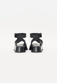 Czarne sandały z grubym słupkowym obcasem, paskami na kostce i odkrytym tyłem. Wykonane z gładkiej sztucznej skóry z minimalistycznymi detalami.