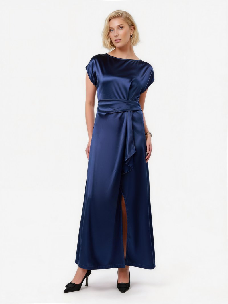 Γυναίκα που στέκεται φορώντας μακρύ φόρεμα από σατέν σε ναυτικό μπλε με κοντά μανίκια, ζώνη στη μέση, σκίσιμο μπροστά και μαύρα μυτερά ψηλοτάκουνα.