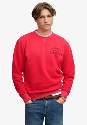 Junger Mann mit hellbraunen Haaren, trägt roten Superdry-Sweatshirt und blaue Jeans, Hände in den Taschen, vor einfachem weißem Hintergrund.