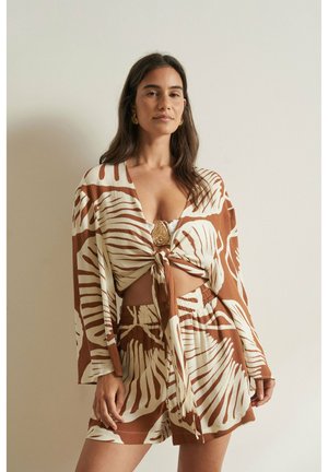 Femme aux longs cheveux foncés portant un haut à nouer devant avec imprimé feuilles marron et beige et un short assorti, debout devant un mur clair uni.
