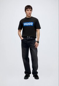 Jonge man staat met handen in zijn zakken, draagt een zwarte HUGO t-shirt, zwarte jeans, zwarte schoenen en een polshorloge tegen een effen achtergrond.