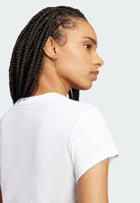 Biela bavlnená tričko s krátkymi rukávmi, s okrúhlym výstrihom a priliehavým strihom. Zobrazuje bočný profil modelky s vrkōčmi.