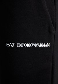 Svart bomullsstoff med trykt hvit tekst: "EA7 EMPORIO ARMANI" med logoakcent, plassert på venstre side.