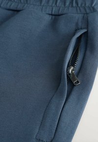 Donkerblauwe joggers met een gladde textuur, voorzien van een ritssluiting aan de zijkant en een elastische tailleband voor een stevige pasvorm.