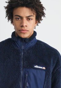 Tamno plavi flis jakna s teksturiranom površinom, visokim ovratnikom i džepovima na patentni zatvarač; sadrži oznaku s logotipom na lijevoj strani prsnog koša.