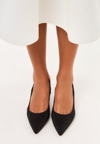 Musta kärkiosa slingback-mokkasiinit, joissa on teksturoitu pinta ja pieniä koristuksia. Muotoilu sisältää matalan korotuksen ja avoimen takapään.