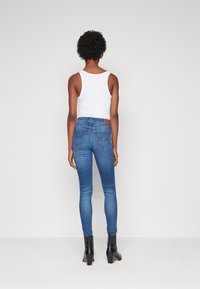 Jean ajustado de mezclilla azul con una cintura de media altura y dos bolsillos traseros. Combinado con botas de tobillo negras. También se ve una camiseta sin mangas blanca.