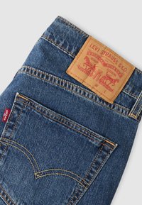Μπλε τζιν παντελόνι Levi's από denim με δερμάτινο ταμπελάκι σε μαύρο δέρμα στη μέση, με κίτρινη ραφή και χαρακτηριστικό σχέδιο στην πίσω τσέπη.